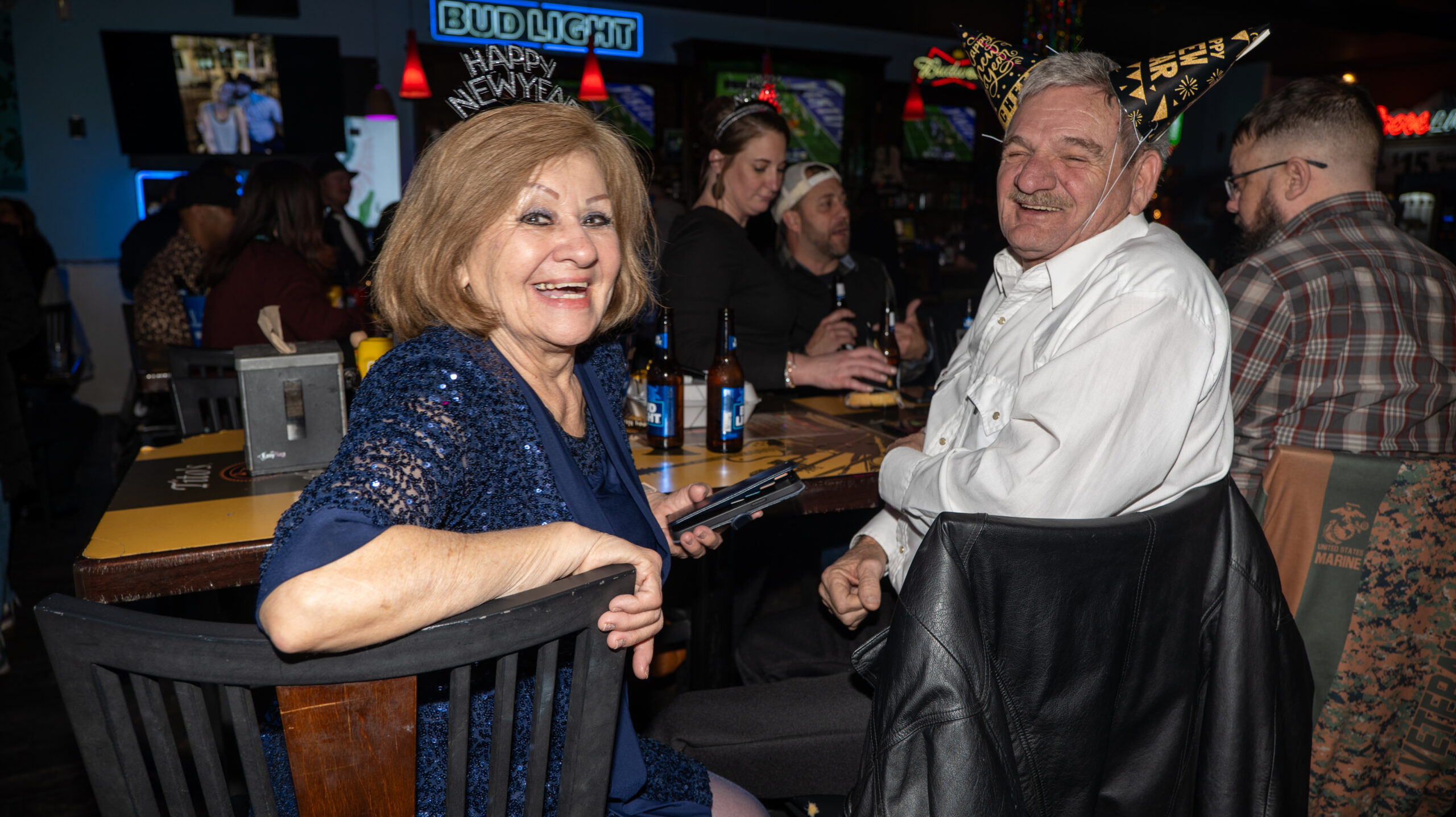 Two senior adults enjoying New Year's Eve at a lively bar, with the woman smiling and holding a smartphone and the man grinning widely, both wearing party hats. In the background, other patrons can be seen interacting and having drinks at the bar, creating a festive atmosphere.